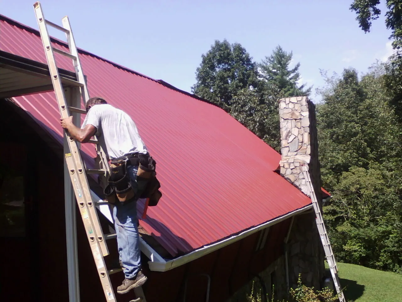 Roofer on a ladder beside a red metal roof and stone chimney — Soffit & Fascia Repair by Roofing Brothers in Martins Creek